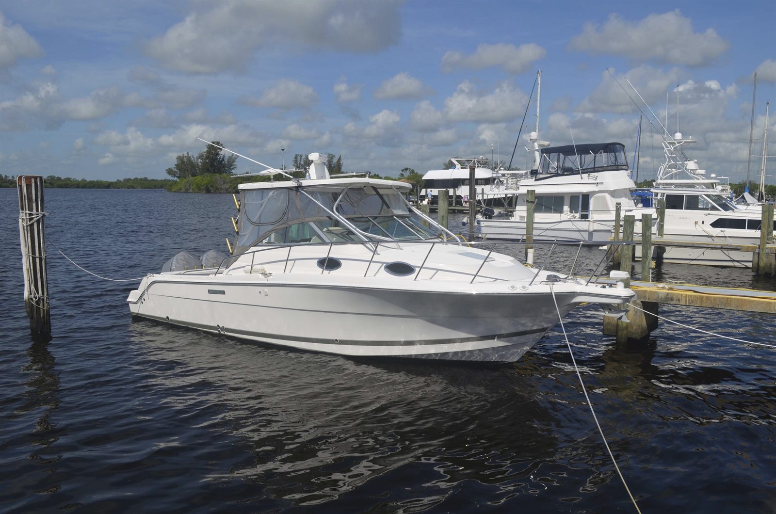 A white motorboat and larger yachts tied up at a marina dock on a wide river